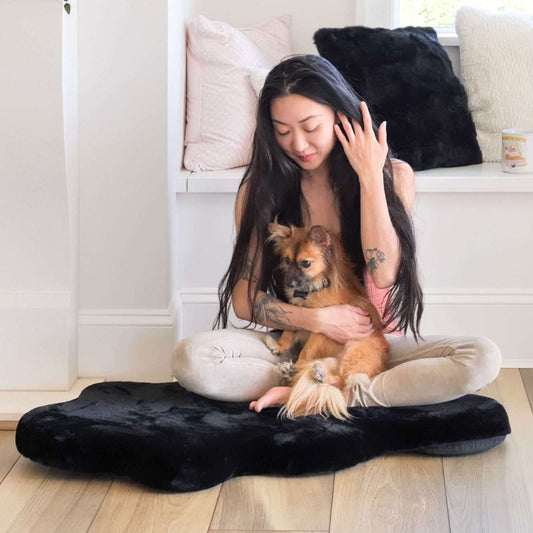 Woman sitting on a large black pet bed with a small dog, surrounded by cushions.