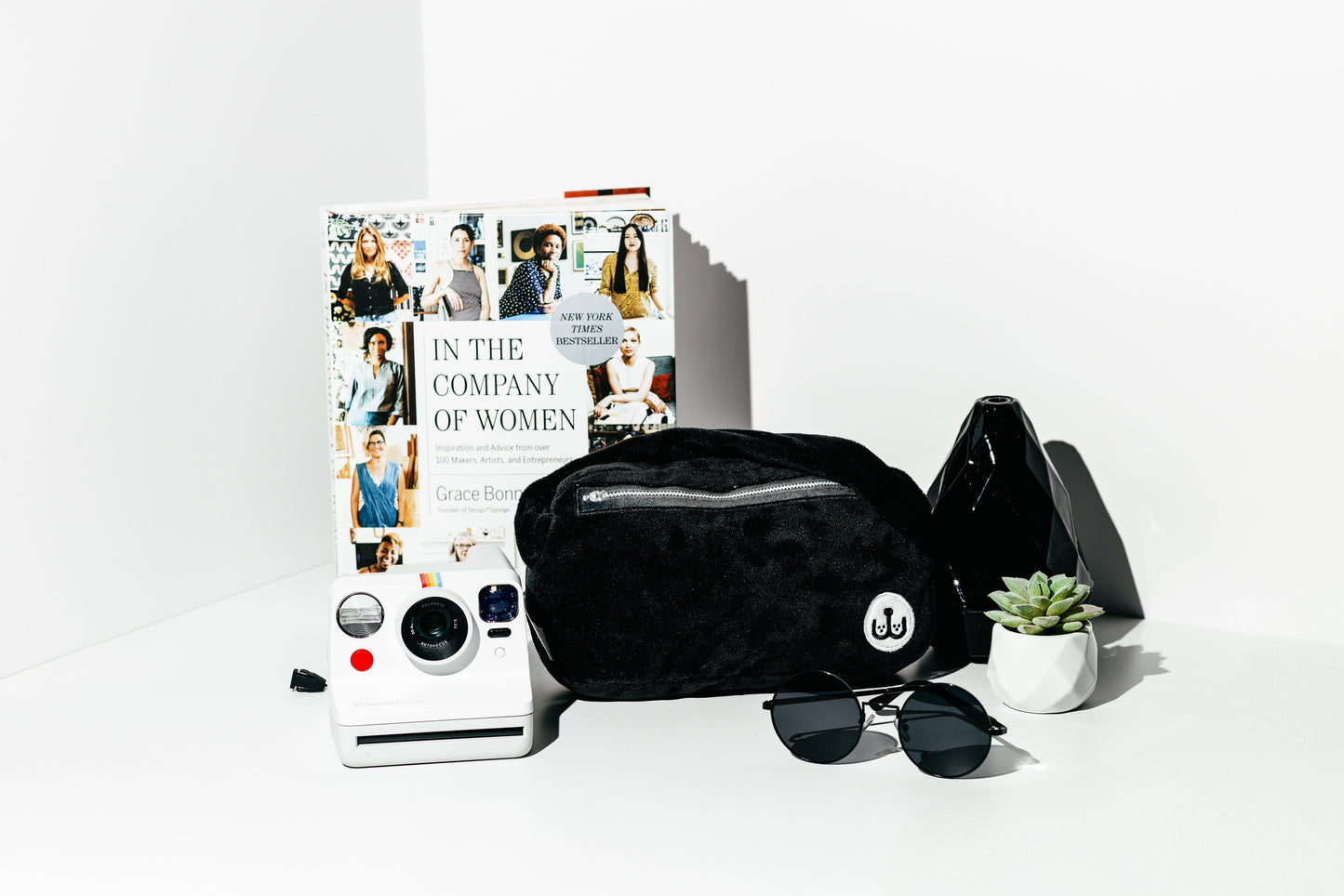 Black quilted bag, sunglasses, and a book titled 'In the Company of Women' on a white surface.