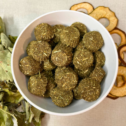 White bowl filled with green herbal balls on a beige background with dried herbs and apples.