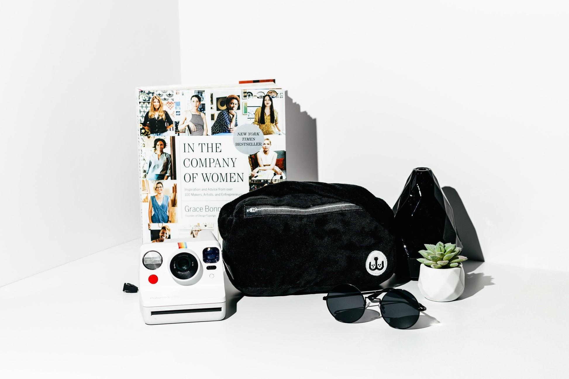 Black quilted bag, sunglasses, and a book titled 'In the Company of Women' on a white surface.