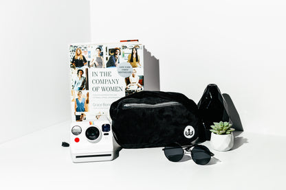Black quilted bag, sunglasses, and a book titled 'In the Company of Women' on a white surface.