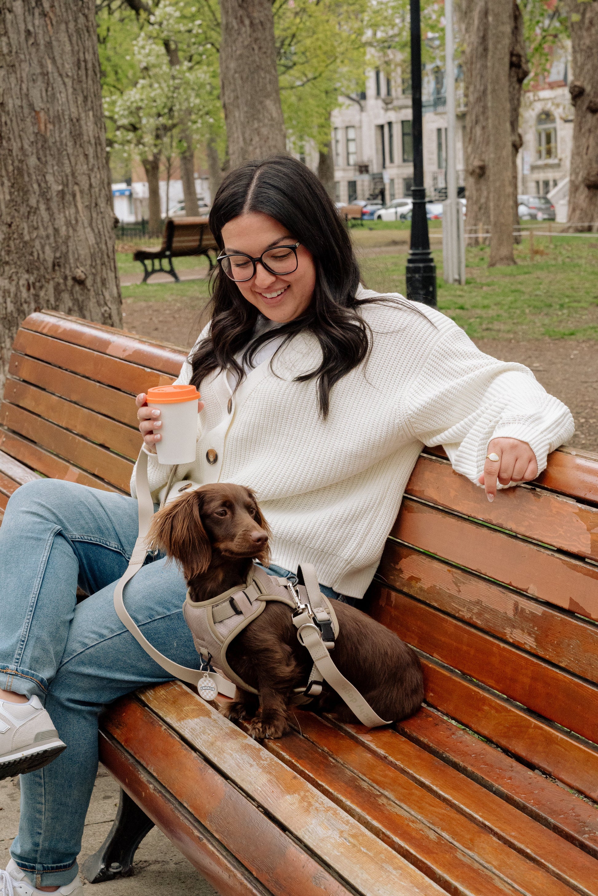 Woman sitting on a park bench with a small dog, holding a cup.