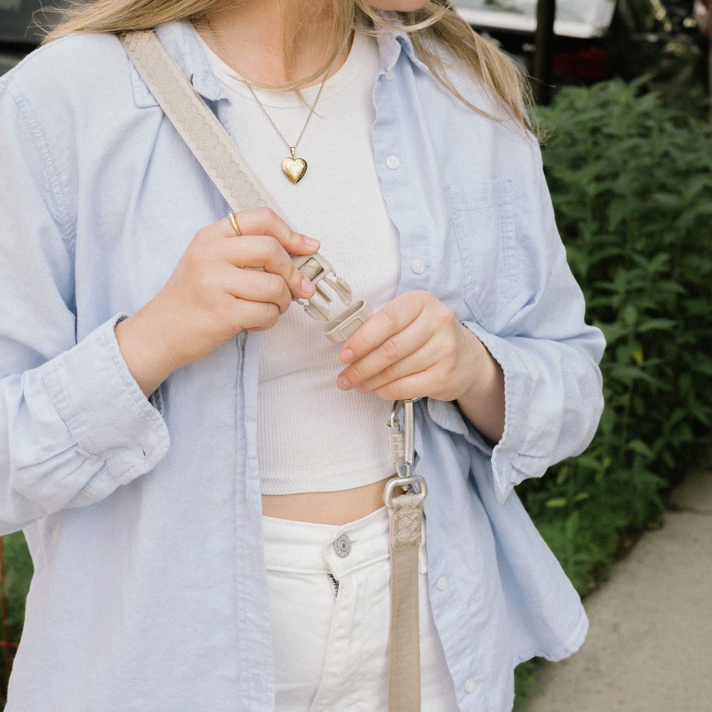 Woman wearing a light blue shirt and white pants with a beige dog walking leash over her shoulder, standing outdoors.